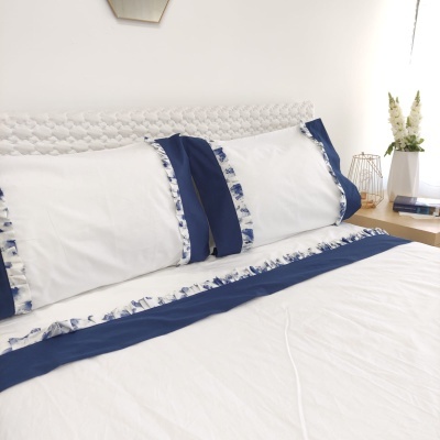 Cama com roupa branca e azul com padrão floral, cabeceira branca e mesa de cabeceira com flores e candeeiro