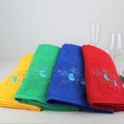 Quatro toalhas coloridas com bordados florais sobre mesa clara com taças e pratos ao fundo.