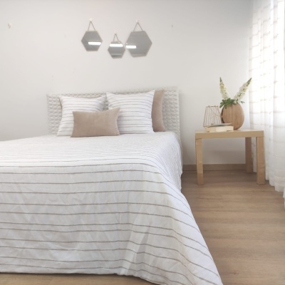 Quarto com cama de colcha branca com riscas finas cinzentas, almofadas brancas e bege, cabeceira estofada branca, mesa de cabeceira de madeira clara com vaso castanho e flores, três espelhos hexagonais na parede branca e piso de madeira clara.