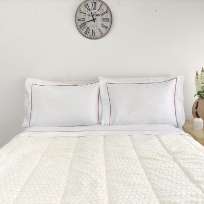 Cama arrumada com almofadas brancas e edredão branco, relógio de parede com números romanos, vaso com flores num ambiente minimalista