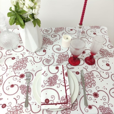 Toalha de mesa branca com padrão vermelho, prato branco com guardanapo, talheres, velas, copos vermelhos e vaso com flores