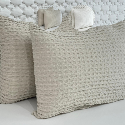Duas almofadas bege claro com textura geométrica sobre superfície branca e cobertor ao fundo