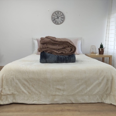 Cama com roupa branca e cobertores castanho e azul empilhados, relógio com números romanos na parede e mesa com vaso e abajur.