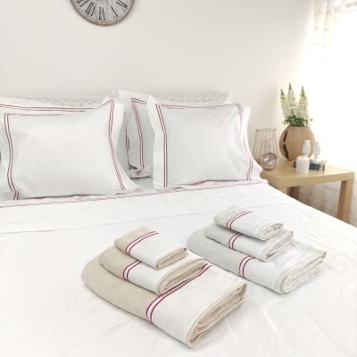 Conjunto de roupa de cama e toalhas brancas com riscas vermelhas sobre cama branca em quarto iluminado.