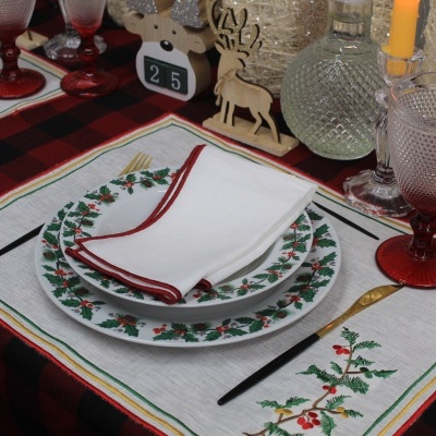 Mesa posta com decoração natalícia, pratos com folhas de azevinho e copos de vidro coloridos.