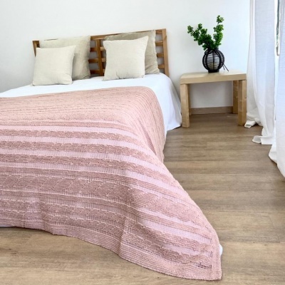 Cobertor rosa claro com padrão listrado sobre cama com almofadas bege e mesa lateral com vaso e planta