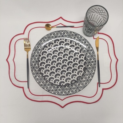 Conjunto de louça preto e branco com talheres dourados e copo de vidro sobre individual branco com bordado vermelho
