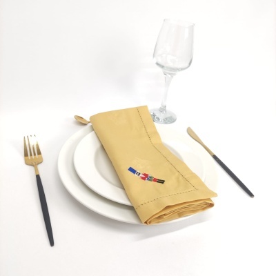 Conjunto de mesa com prato branco, talheres preto e dourado, guardanapo amarelo bordado e copo de vidro