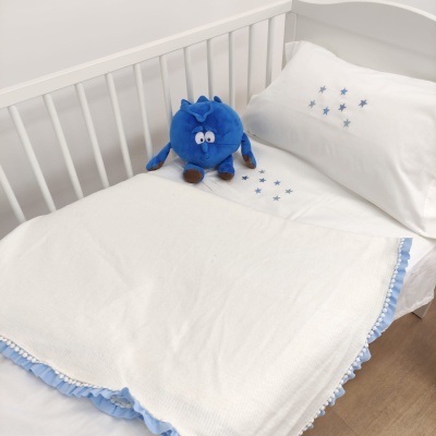 Cama de bebé com lençóis brancos e estrelas azuis, manta branca com bordado azul e peluche azul num berço branco