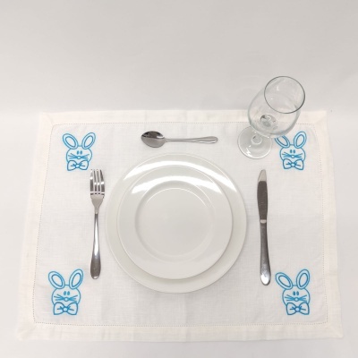 Jogo americano branco com bordado azul de coelhos, pratos e talheres sobre fundo branco