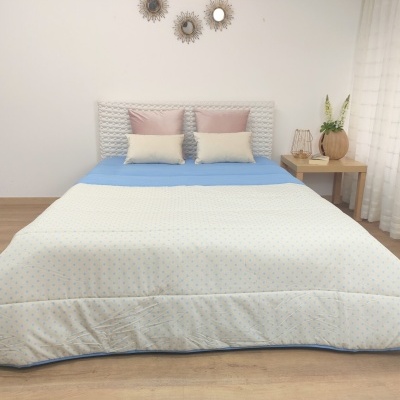Cama arrumada com colcha azul clara e edredão branco com padrão floral, almofadas rosa e bege, mesa lateral com vaso e decoração