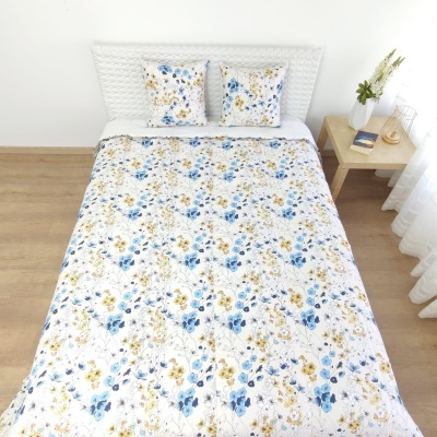 Edredão e fronhas florais azuis e amarelas em cama branca num quarto com chão de madeira e mesa de cabeceira.