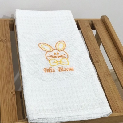 Toalha branca com bordado de coelho e texto 'Feliz Páscoa' sobre suporte de madeira