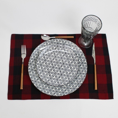 Conjunto de jantar com pratos brancos e pretos, talheres com cabo de madeira, copo transparente em tecido xadrez vermelho e preto.