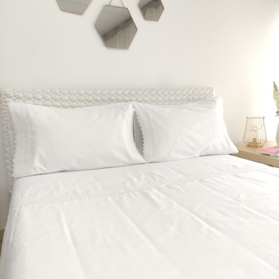 Jogo de cama branco bordado numa cama com cabeceira acolchoada e mesa de cabeceira com candeeiro e vaso