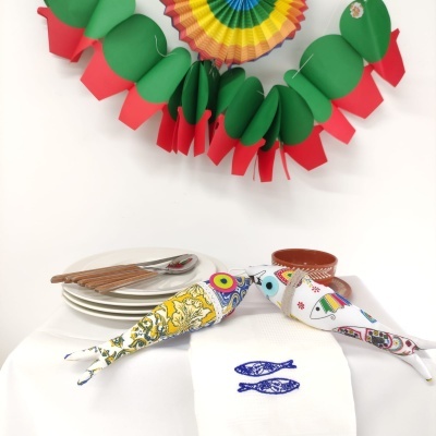 Mesa com toalha branca, dois peixes decorativos de tecido colorido, pratos, talheres e panela, decoração verde, vermelha e roseta colorida pendente