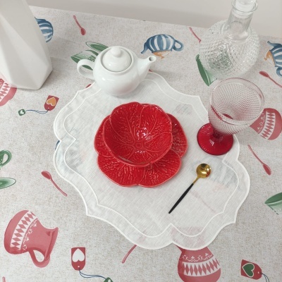 Conjunto de chá com bule branco, pratos vermelhos em forma de flor, taça transparente com base vermelha, colher pequena dourada, jarro de vidro e toalha colorida com padrões de chá