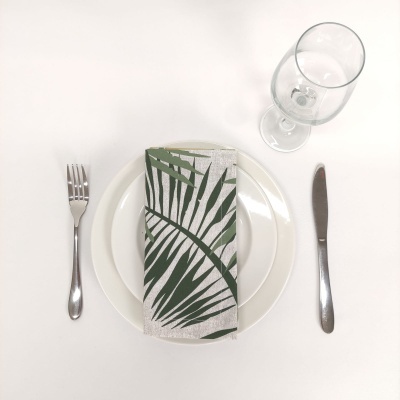 Conjunto de mesa posto com prato branco, guardanapo com folhas verdes, talheres prateados e copo de vidro.