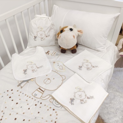 Conjunto de roupa e acessórios para bebé com tema de ursos, em branco com bordados castanhos e cinzentos, num berço branco.