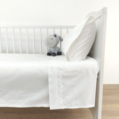 Cama branca com lençóis brancos detalhados, almofada e peluche cinzento junto à cabeceira