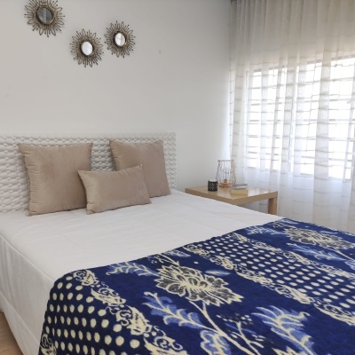 Quarto com cama branca, manta azul com padrão, almofadas castanhas, espelhos redondos, mesa de cabeceira e cortinas brancas