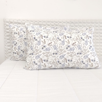 Dois almofadas com padrão floral azul e castanho sobre cama com roupa branca e cabeceira acolchoada
