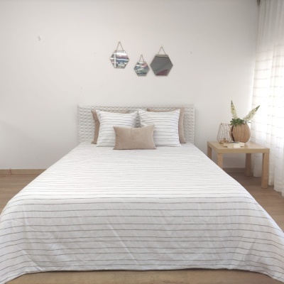 Cama de casal com roupa de cama branca de riscas finas cinza e almofadas bege, mesa de cabeceira com vaso e candeeiro