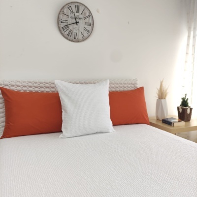 Cama com roupa branca e almofadas laranja e branca, relógio na parede e mesa de cabeceira com decoração