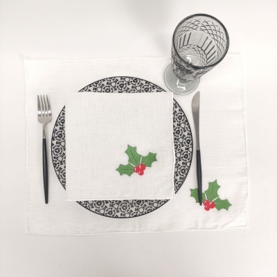 Conjunto de jantar com toalha branca com decoração de folhas verdes e bagas vermelhas, prato com padrão preto, talheres e copo transparente