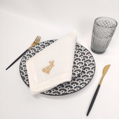 Mesa posta com prato decorado, guardanapo branco bordado, talheres dourados e pretos e copo de vidro cinza