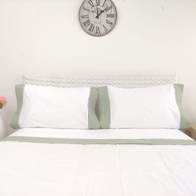 Cama arrumada com almofadas brancas e detalhes verdes, cabeceira trançada branca e relógio de parede com números romanos