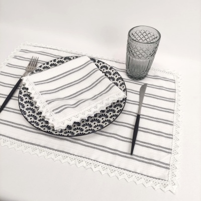 Lugar de mesa com prato preto e branco, guardanapo com riscas, garfo e faca com cabo preto, copo cinzento texturizado e toalha branca com riscas cinzentas