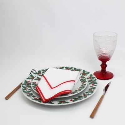 Conjunto de pratos decorados com azevinho, guardanapo branco com bordas vermelhas, talheres de cabo madeira e taça com base vermelha.