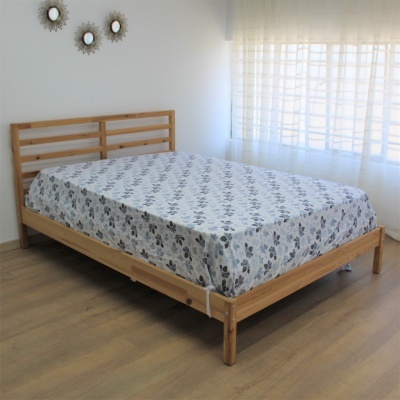 Cama de madeira clara com lençol cinza com padrão floral em quarto iluminado