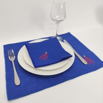 Mesa posta com guardanapo e toalhete azuis bordados, prato branco, talheres metálicos e copo de vidro