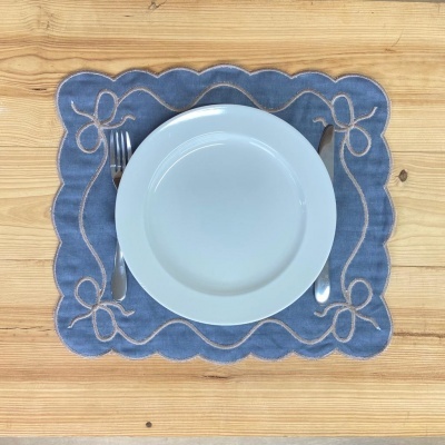 Lugar americano azul com laços bordados, prato branco, garfo e faca sobre mesa de madeira.