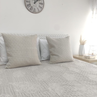 Cama com almofadas bege, relógio de parede, cortina branca e mesa de cabeceira clara com decoração