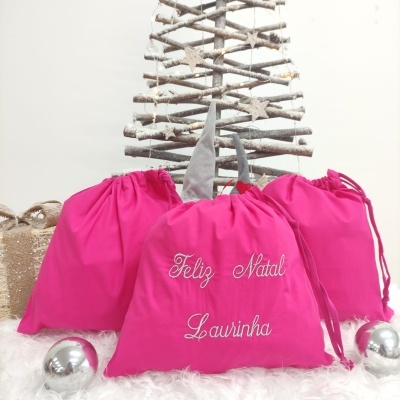 Três sacos rosa com cordão e bordado 'Feliz Natal Laurinha' em cenário natalício com árvore de madeira e bolas prateadas