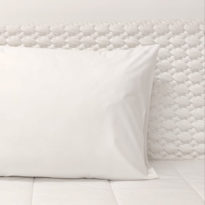 Travesseiro branco com fronha lisa sobre colchão branco acolchoado, encostado em cabeceira branca texturizada.