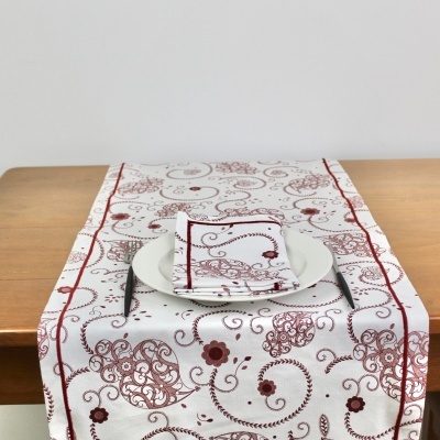 Toalha de mesa branca com padrão ornamental vermelho e castanho numa mesa de madeira com prato, guardanapo e talheres