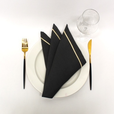 Conjunto de prato branco, talheres pretos e dourados, guardanapos pretos com riscas douradas e copo de vidro