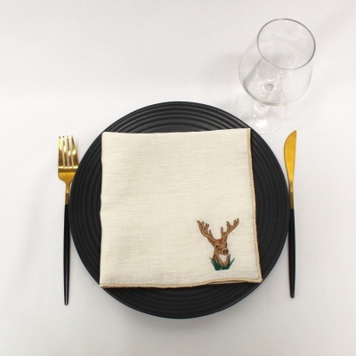 Conjunto de louça com prato preto, guardanapo branco bordado com veado, talheres dourados com cabo preto e copo de vidro.