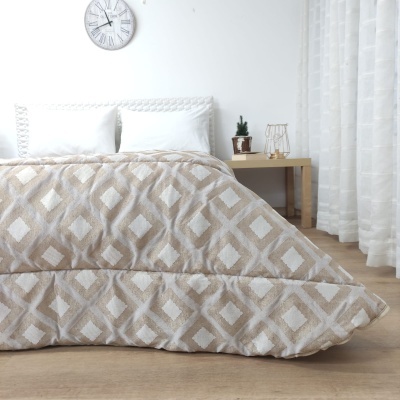 Edredão bege e branco com padrão de losangos numa cama minimalista num quarto com mesa de cabeceira e cortinas brancas.