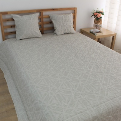 colcha cinzenta clara com padrão geométrico, duas almofadas iguais, cama de madeira clara e mesa com vaso de flores