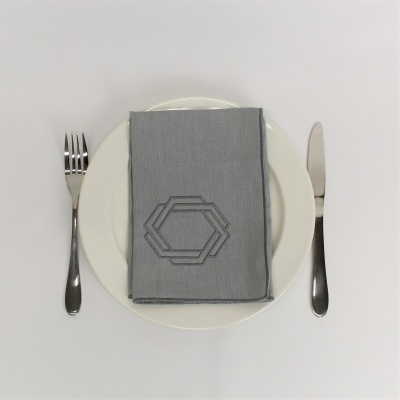 Guardanapo cinzento com padrão geométrico bordado em prato branco com talheres à esquerda e direita.