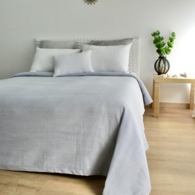 Cama de casal com colcha e almofadas em tons cinzentos e mesa de cabeceira com vaso na parede branca