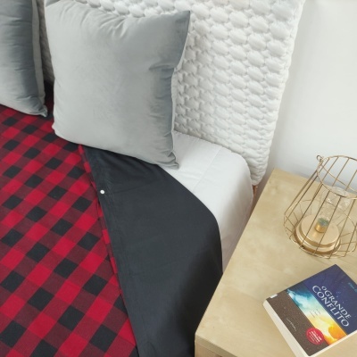 Cama com almofada cinza, colcha branca texturizada, manta xadrez vermelha e preta, mesa de cabeceira com vela e livro