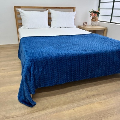 Cobertor azul texturizado sobre cama com lençol branco e almofadas brancas, em quarto com chão e móveis claros e janela preta.