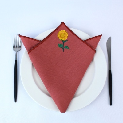 Guardanapo terracota com bordado de girassol sobre prato branco, garfo e faca com cabos pretos