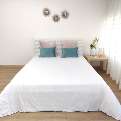 Cama com colcha branca, almofadas rosa e azul, espelhos redondos na parede, mesa de cabeceira com flores e abajur.